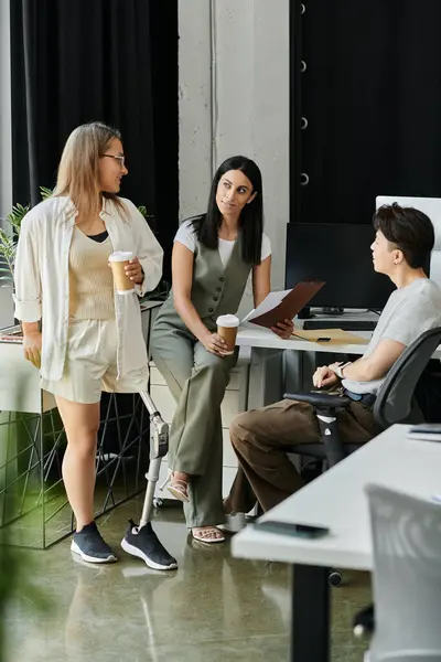 Three coworkers stand and sit in a modern office, talking and drinking coffee. — Stock Photo