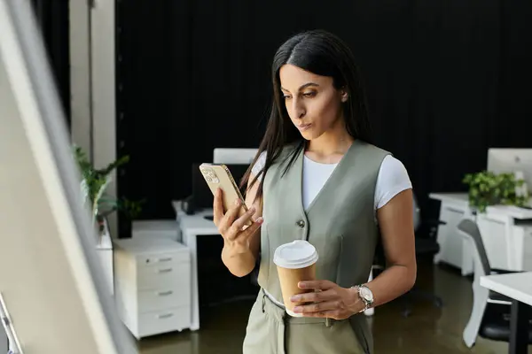 A woman in a green vest checks her phone while holding a coffee cup. — Stock Photo