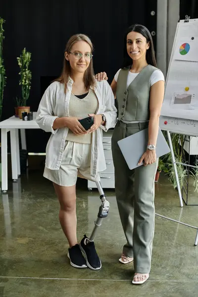 Due donne in piedi in un ambiente ufficio, una con una gamba protesica, sorridente e guardando la fotocamera. — Stock Photo