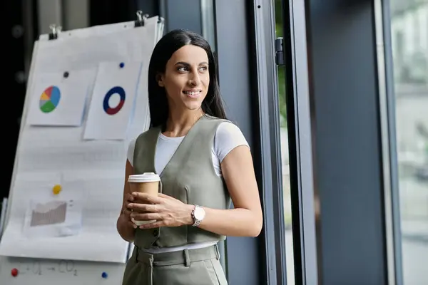 Una donna si trova in un ufficio moderno, tenendo un caffè e guardando fuori da una finestra. — Foto stock