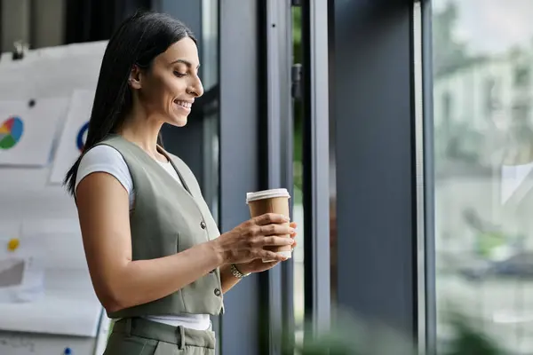 A woman in a green vest stands by a window, holding a coffee cup and looking out. — Stock Photo