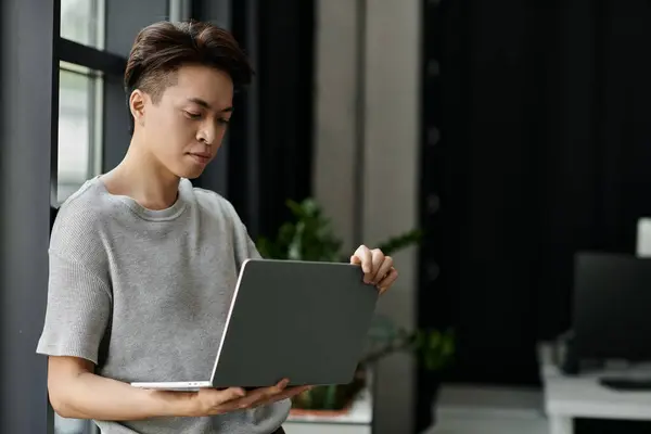 Ein junger Mann steht an einem Fenster in einem Büro und konzentriert sich auf seinen Laptop. — Stockfoto
