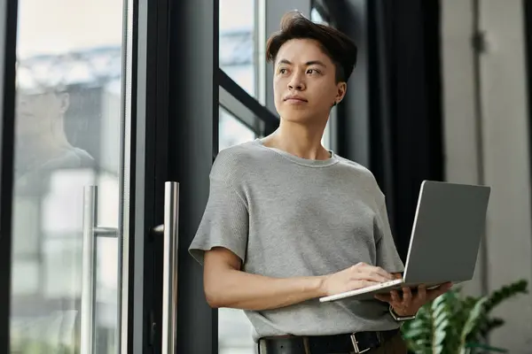 A young man stands by a window, laptop in hand, looking thoughtfully out at the city. — Stock Photo