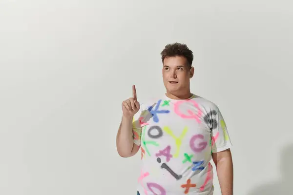 A queer person wearing a white shirt with colorful graffiti points up with their finger. — Stock Photo