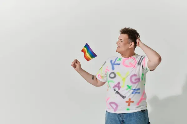 Una persona queer con una camicia colorata regge una bandiera arcobaleno e sorride. — Foto stock