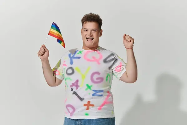 A queer person with a colorful shirt proudly holds a rainbow flag and raises their fist in the air. — Stock Photo