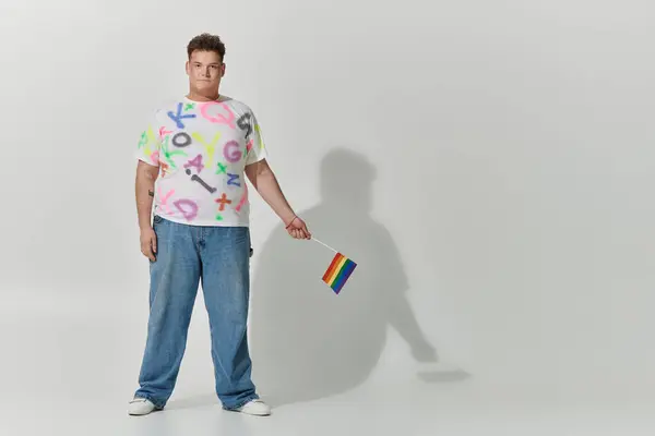 A queer person in a colorful shirt and jeans stands holding a rainbow flag. — Stock Photo