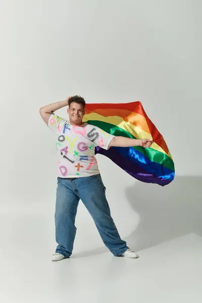 A queer person in a white shirt with colorful letters and blue jeans poses with a rainbow flag. — Stock Photo