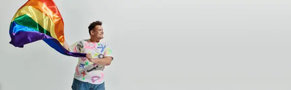 Una persona queer in abiti colorati tiene una bandiera arcobaleno, sorridente e guardando al lato. — Foto stock