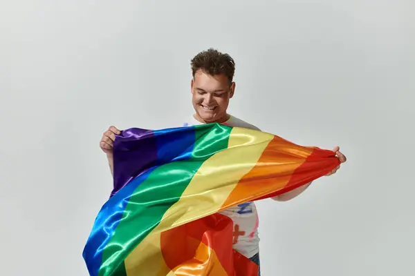 Una persona queer sonríe mientras sostiene una bandera de arco iris frente a un fondo blanco. — Stock Photo