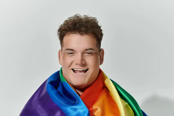 Una persona queer drappeggiato in una bandiera arcobaleno sorride ampiamente. — Foto stock