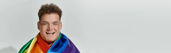 A young man smiles brightly, draped in a vibrant rainbow flag. — Stock Photo