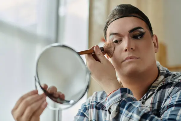 Una persona queer aplica maquillaje, cuidadosamente usando un cepillo delante de un espejo. - foto de stock
