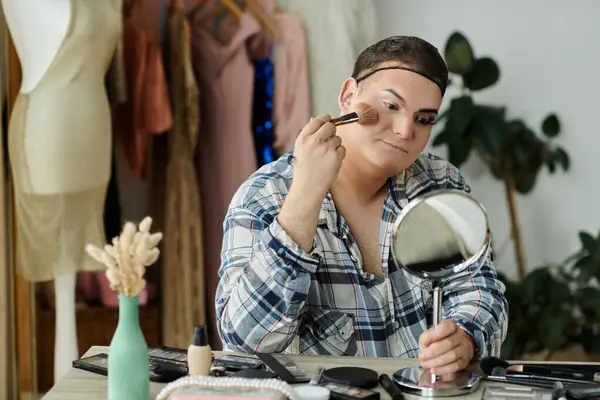 A queer person applies makeup in a room with a mirror and makeup brushes. — Stock Photo