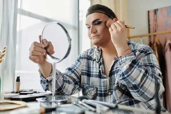 A queer person in a plaid shirt applies makeup while looking in a mirror. — Stock Photo