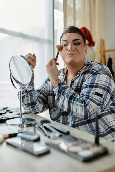 A queer person in a plaid shirt applies makeup with a brush while looking in a mirror. — Stock Photo