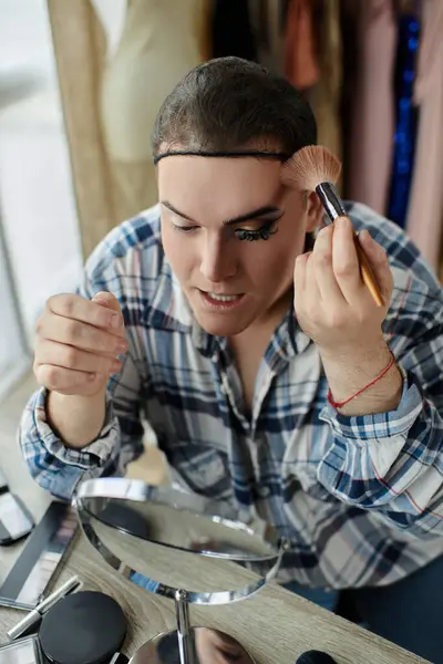 A queer person applies makeup while looking in a mirror, wearing a stylish plaid shirt. — Stock Photo