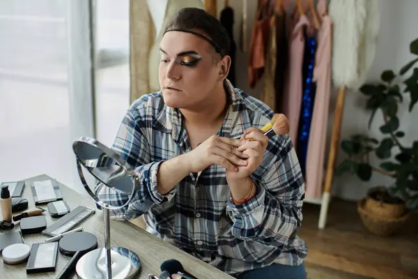 A queer person applies makeup in front of a mirror, surrounded by their beauty tools. — Stock Photo