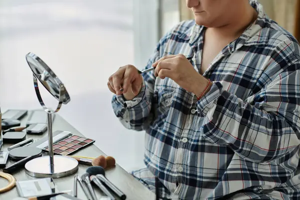 Une personne queer applique le maquillage devant un miroir, entouré de leurs outils de beauté. — Photo de stock