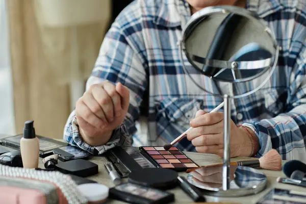 A queer person applies eyeshadow while looking in a mirror, surrounded by makeup products. — Stock Photo