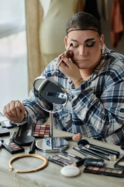 A queer person applies eye makeup in front of a mirror, surrounded by various makeup tools and products. — Stock Photo