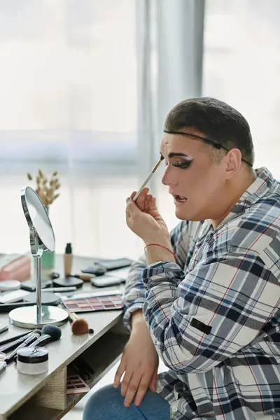 Una persona queer aplica maquillaje delante de un espejo, usando una camisa a cuadros y jeans. — Stock Photo