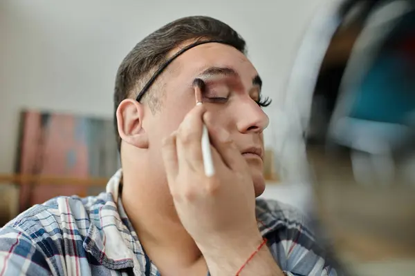 A queer person applies makeup to their eye with a brush, focusing on their reflection. — Stock Photo