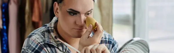 Una persona queer aplica maquillaje con una esponja mientras se mira atentamente en un espejo. - foto de stock