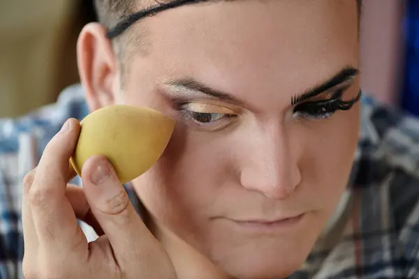 Una persona queer aplica maquillaje con una esponja, centrándose en su área de los ojos. - foto de stock