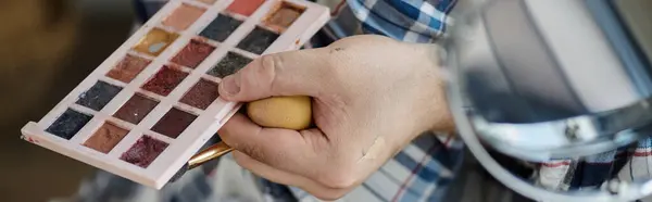 La mano di una persona queer tiene una spugna e un pennello per il trucco, con una tavolozza di ombretti sullo sfondo. — Foto stock