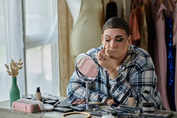 A queer person applies makeup while looking in a mirror, surrounded by makeup supplies. — Stock Photo