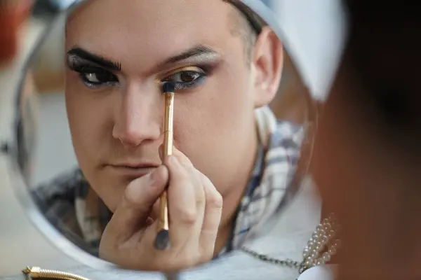 A young queer person applies makeup while looking in the mirror. — Stock Photo