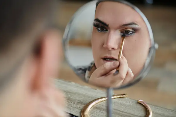 A queer person applying makeup while looking at their reflection in a mirror. — Stock Photo