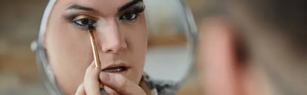 A close-up of a queer person applying eyeshadow with a brush, looking at themselves in the mirror. — Stock Photo