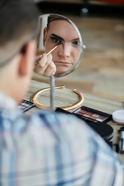 A queer person applies eyeliner with a brush, looking intently in a mirror. — Stock Photo