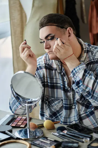 A queer person wearing a plaid shirt applies eyeliner in front of a mirror. — Stock Photo