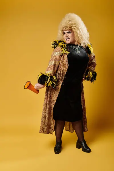 A drag queen in stylish clothing holds a megaphone against a yellow background. — Stock Photo