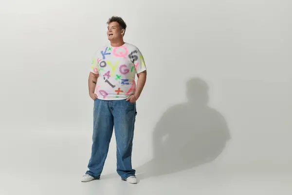 A queer person in white shirt and jeans with colorful letters stands confidently against white backdrop — Stock Photo