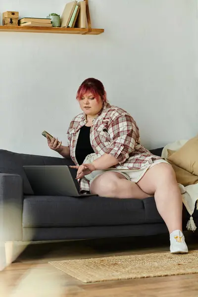 Женщина plus size в рубашке сидит на диване и работает на ноутбуке. — стоковое фото