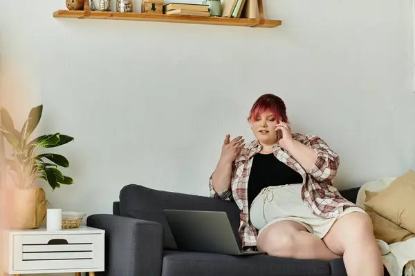 Uma mulher senta-se em um sofá, falando em um telefone e trabalhando em um laptop. — Fotografia de Stock