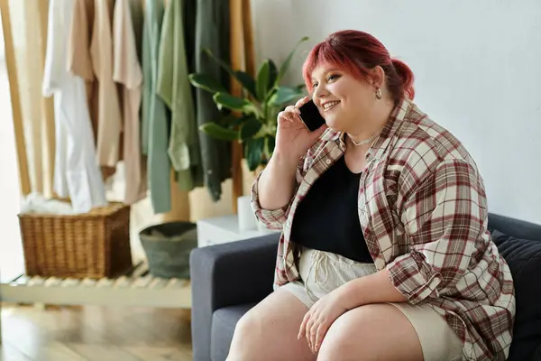 Una donna plus size siede su un divano, sorridente mentre parla al telefono. — Foto stock