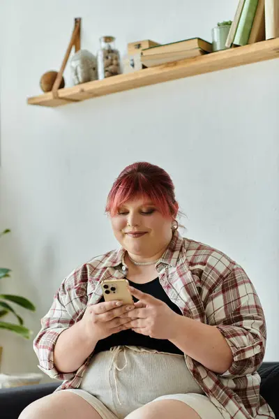 Pink-haired plus-size woman in plaid shirt smiles on her phone at home — Stock Photo