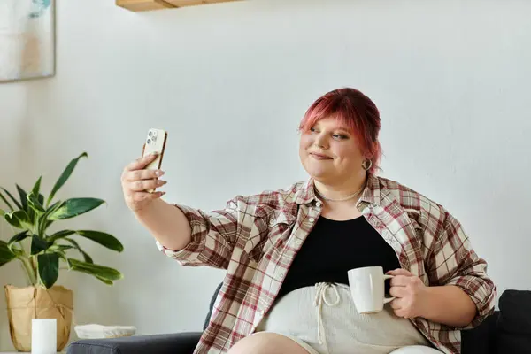 Una mujer se sienta en un sofá, sonriendo mientras se toma una selfie con su teléfono. - foto de stock