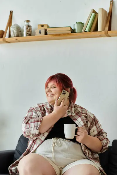 Una mujer con una camisa a cuadros se sienta en un sofá en casa, sosteniendo un teléfono en la oreja y una taza en la mano. — Stock Photo