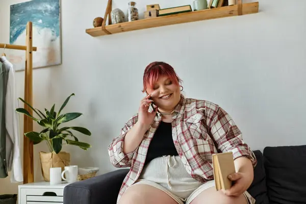 Eine Plus-Size-Frau im karierten Hemd sitzt auf ihrer Couch und plaudert fröhlich am Telefon.. — Stockfoto