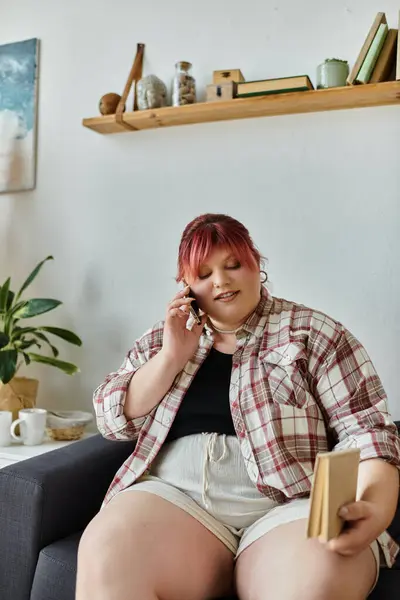 Una mujer se sienta en un sofá, hablando por teléfono y sosteniendo un libro. - foto de stock
