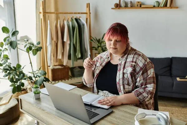 Una donna in camicia a quadri si siede a una scrivania con un computer portatile, un notebook e una penna, lavorando a casa.. — Stock Photo