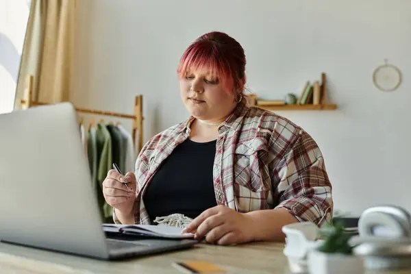 Una donna con i capelli rosa siede a una scrivania, scrivendo in un taccuino con un computer portatile aperto di fronte a lei. — Foto stock