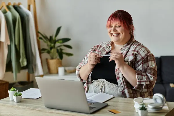 Женщина plus size с рыжими волосами сидит за столом и улыбается, работая над ноутбуком. — стоковое фото