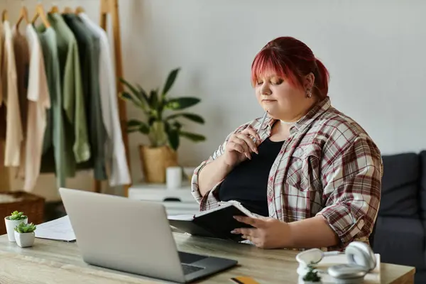 Curvy woman with red hair sitting at home office desk, gazing down at notebook — Stock Photo